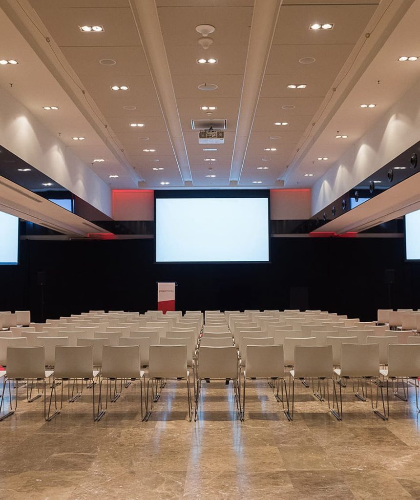 Vista frontal de una sala de eventos, con las sillas, el escenario con atril y la pantalla